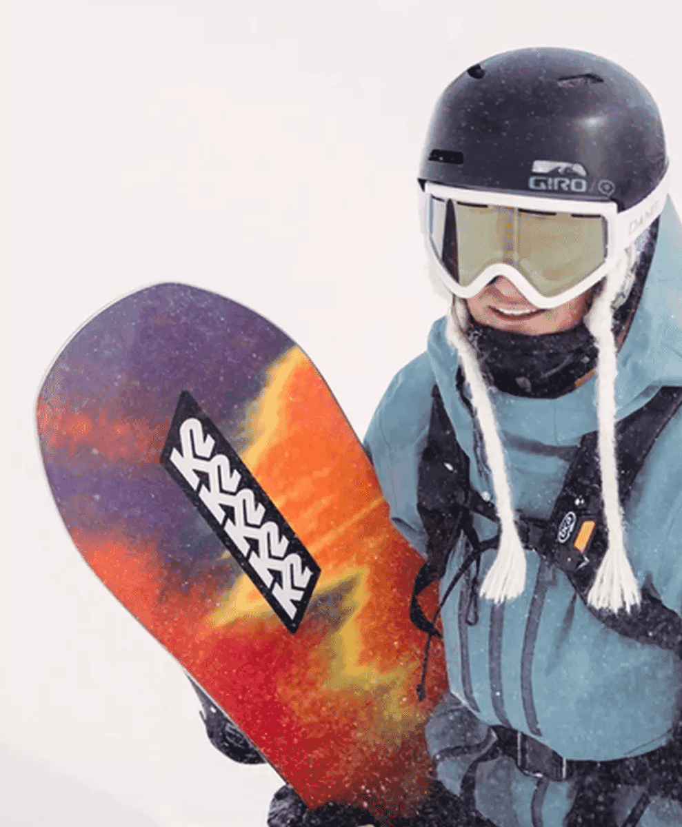 A smiling snowboarder in a teal jacket holding a colorful snowboard in the snow.