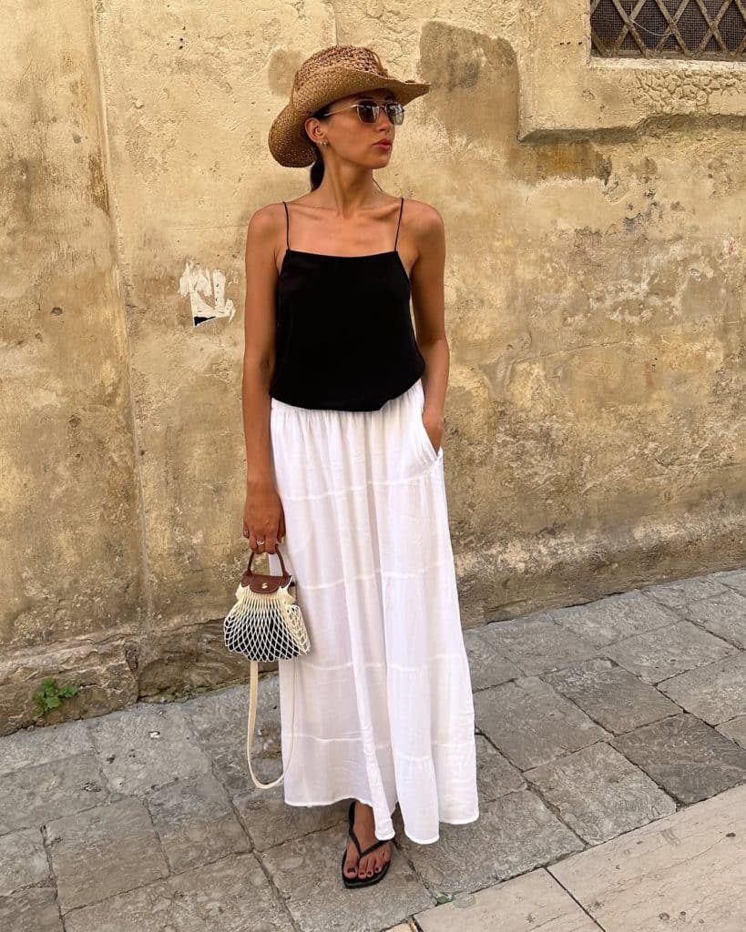 A woman in a black spaghetti strap top, white skirt, and straw hat on a stone street.