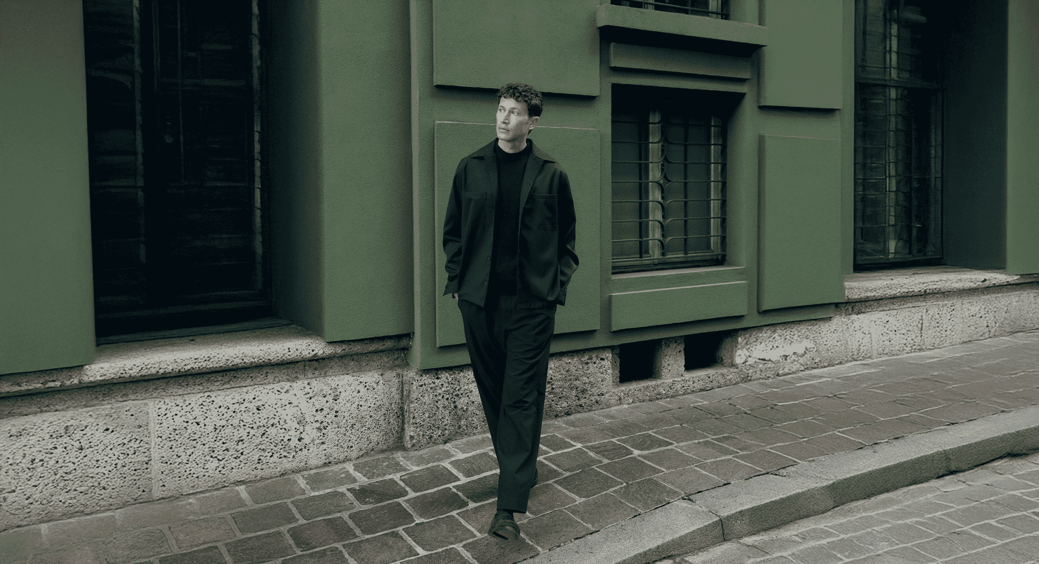 Full shot of a man in an all-black minimalist jacket and trousers on a cobblestone street.
