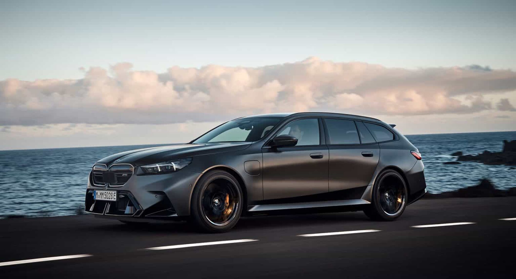 Matte dark gray BMW M5 Touring driving on a coastal road during dusk with the ocean in the background.