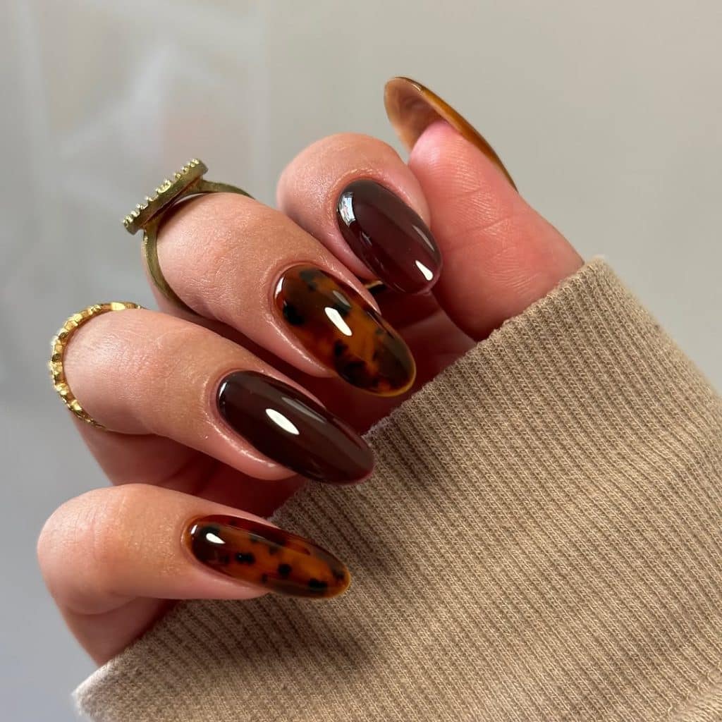 Hand with tortoiseshell nail art and gold rings emerging from a beige ribbed sweater sleeve.