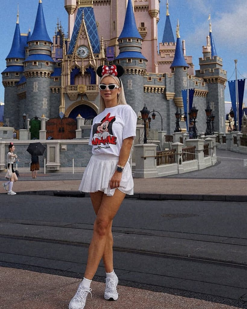 A woman wearing a white Minnie Mouse T-shirt, pleated skirt, and Mickey ears standing before the castle.