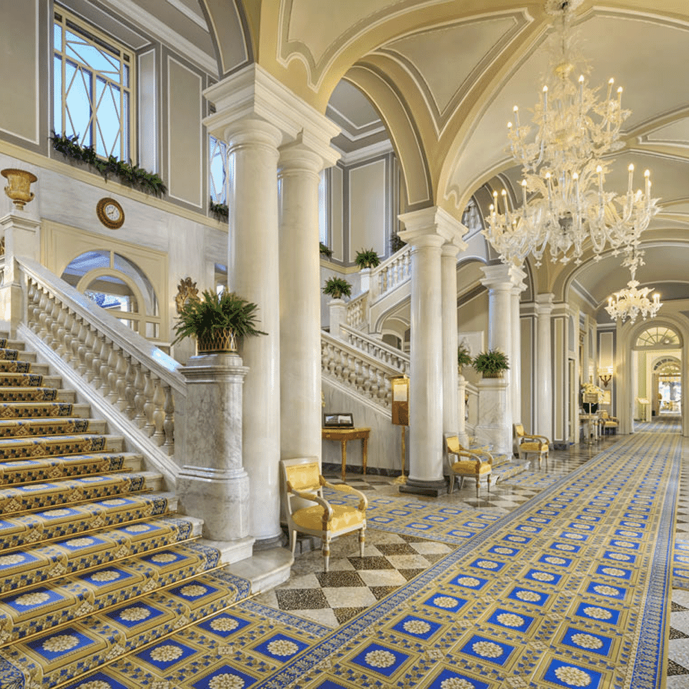 Opulent hotel lobby with marble staircases, crystal chandeliers, and ornate blue-and-gold carpets.