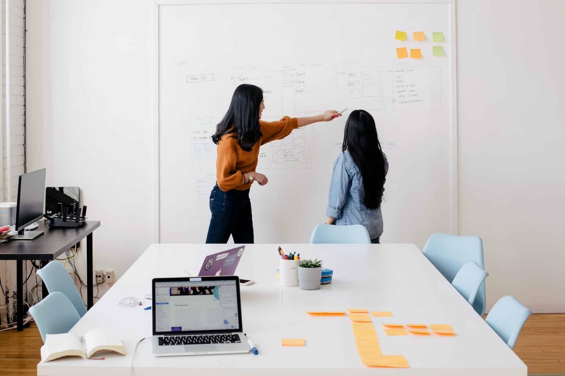 Two women, one in an orange sweater and one in a denim jacket, working at a whiteboard in a modern office.