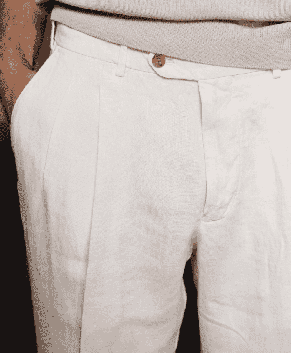 Close-up of white pleated linen trousers showing the fine fabric grain and tailored details.