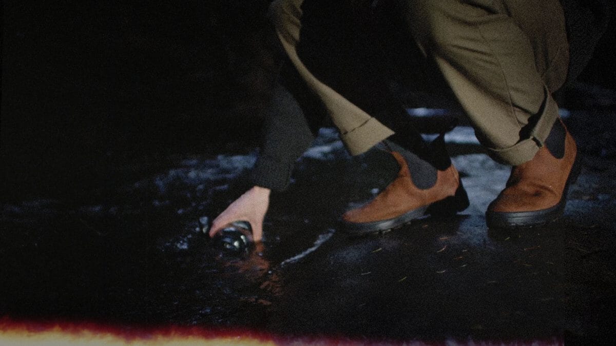 Person in cargo pants and suede Chelsea boots crouching on a wet, reflective urban surface at night.