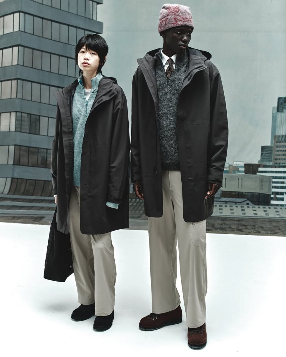 Two models posing in dark coats and beanies with a city skyline in the background.