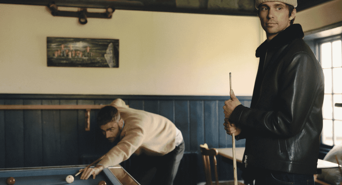 Two men in a pool hall, one wearing a black leather bomber jacket and the other a cream knit sweater.
