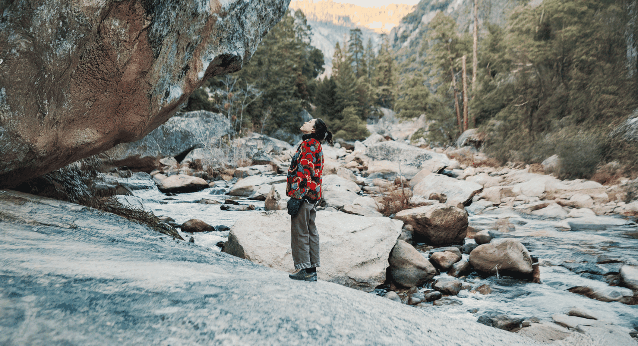 A person on a rocky riverbank wearing a red patterned sweater and wide-leg pants gazing upward.