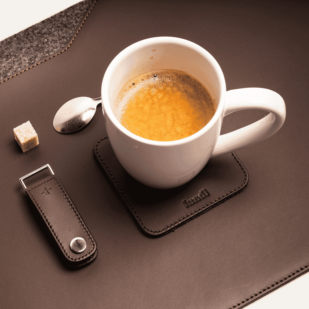 A white ceramic coffee cup resting on a branded brown leather coaster next to lifestyle accessories.