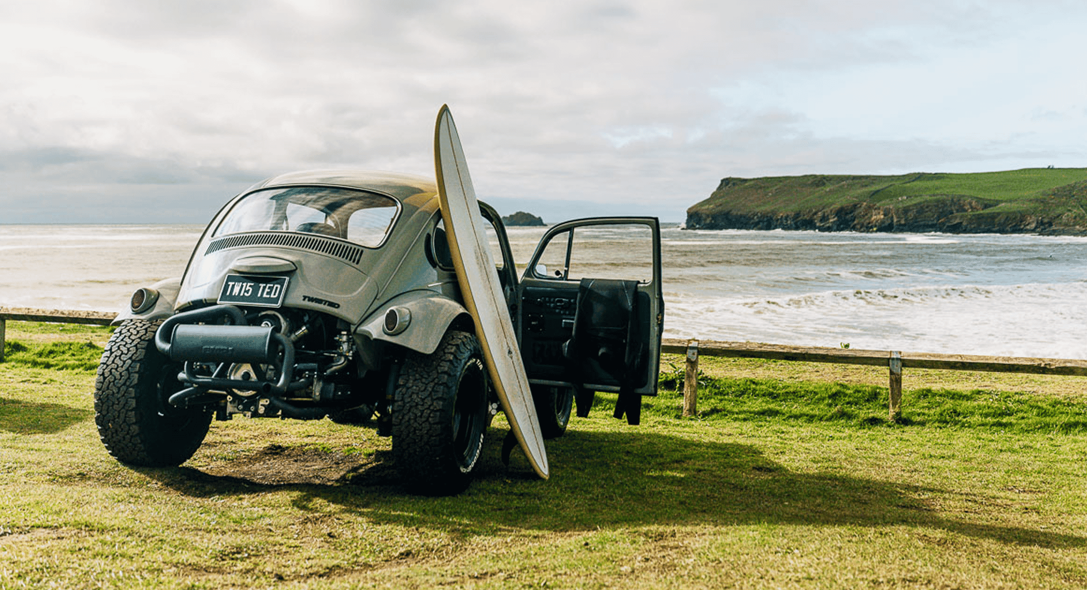 Customized vintage Beetle with surfboard on a grassy cliff.