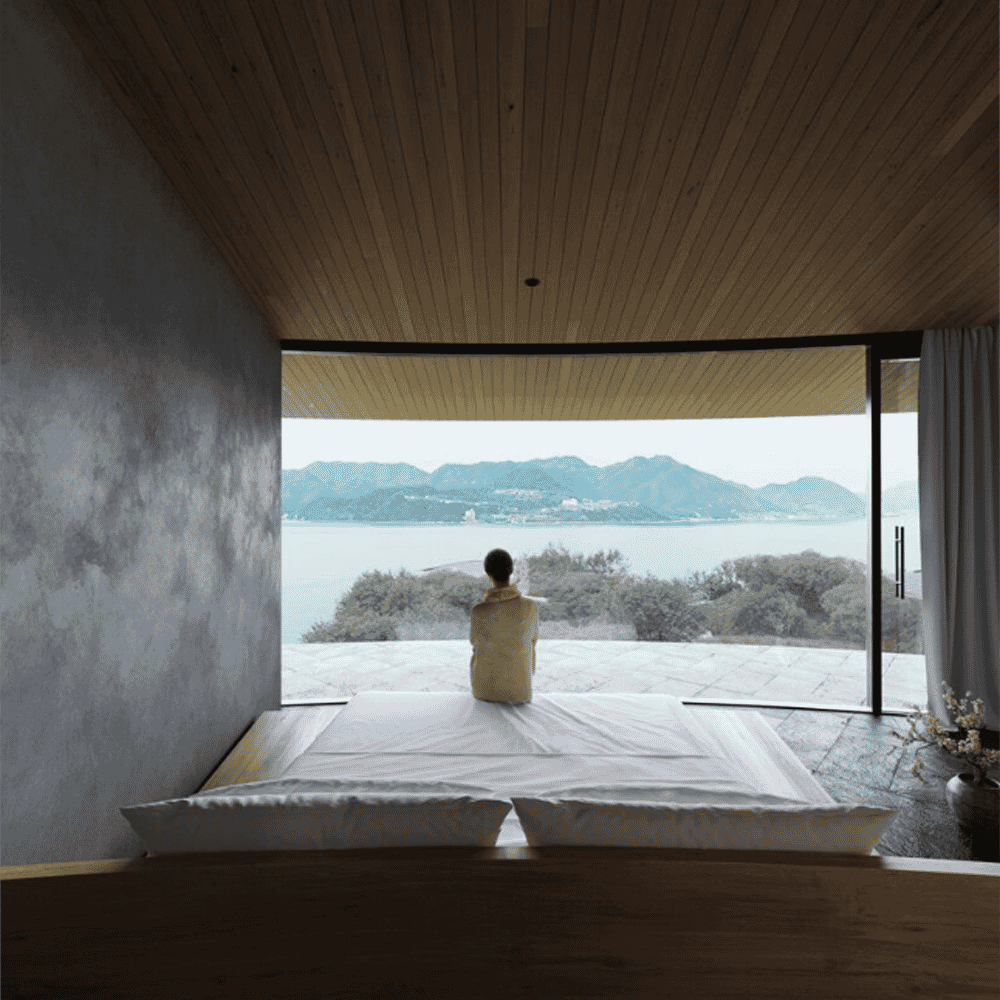 A person in a neutral-toned hooded robe sitting in a minimalist room looking out at the sea through a large window.