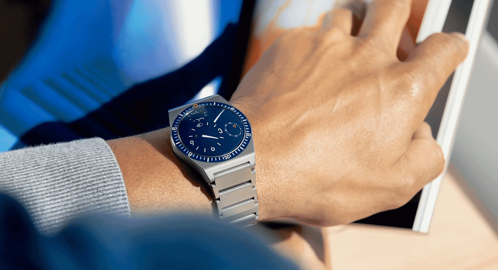 A man's wrist wearing a blue dial watch paired with a gray ribbed sweater.