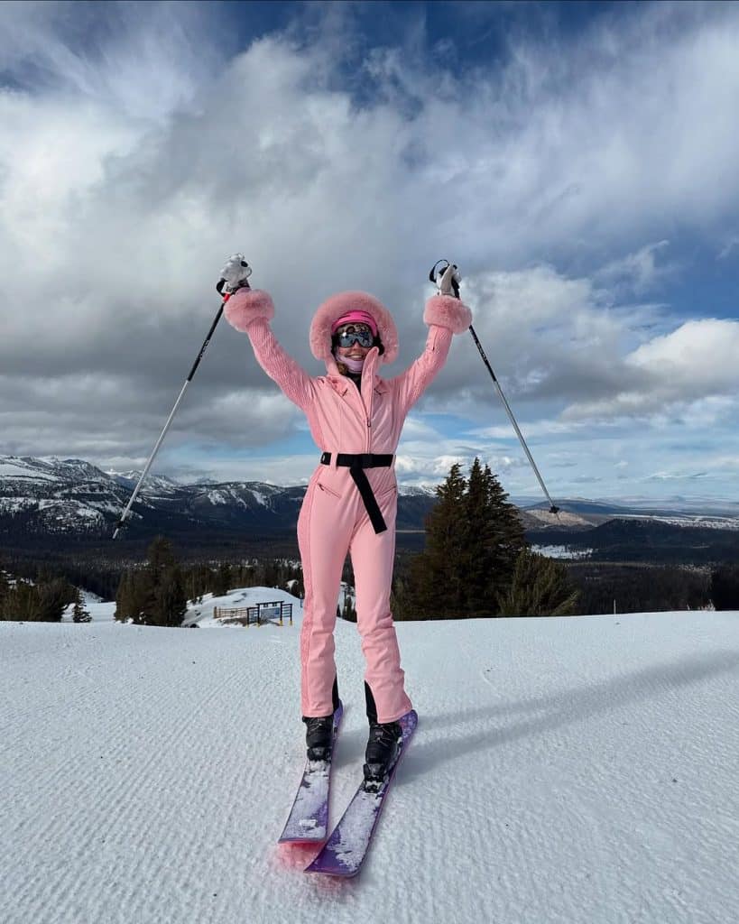 Skier in a vibrant pink fur-lined suit with a black belt celebrating on the slopes.
