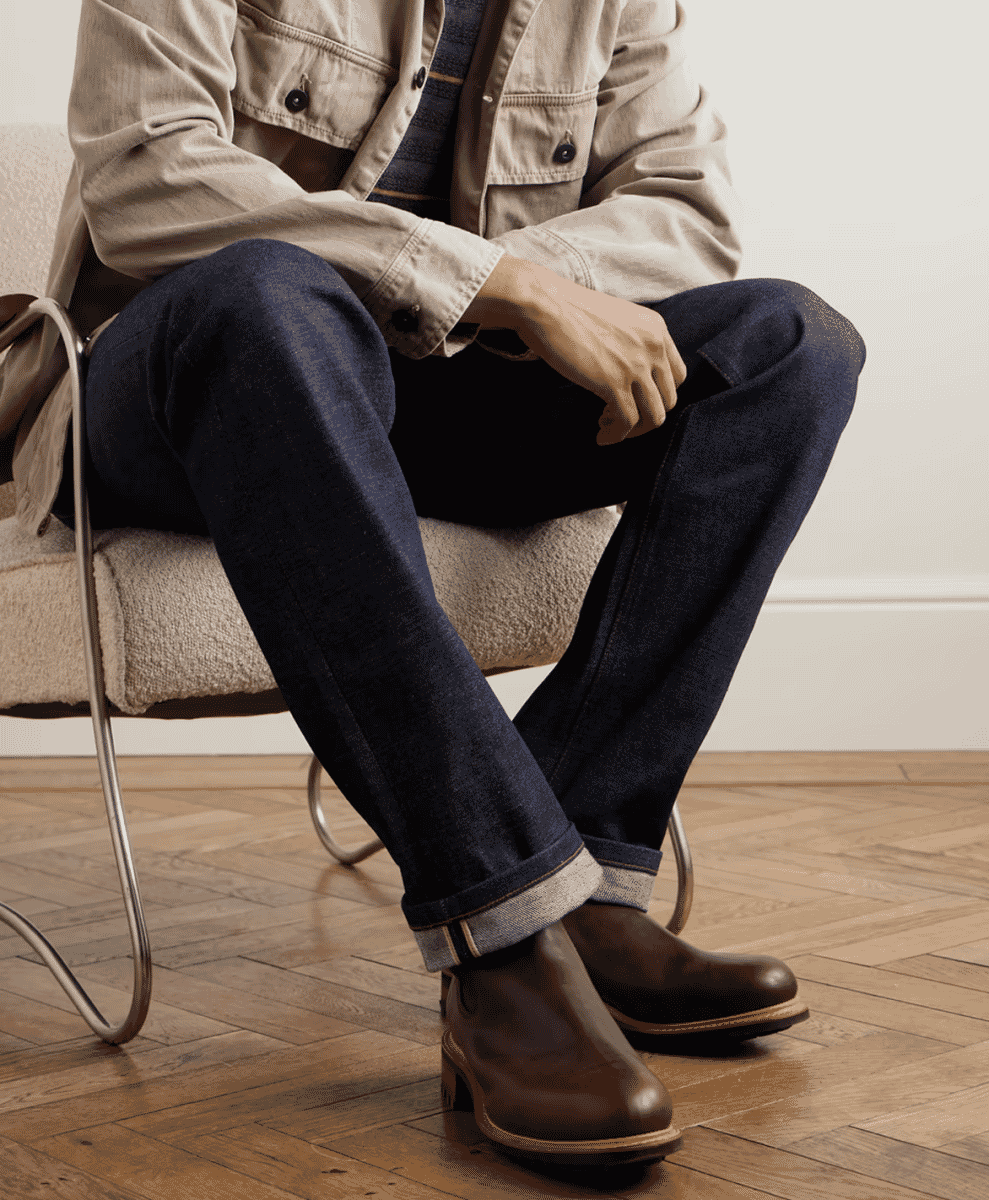 A man in a utility jacket and cuffed denim jeans wearing brown leather Chelsea boots.