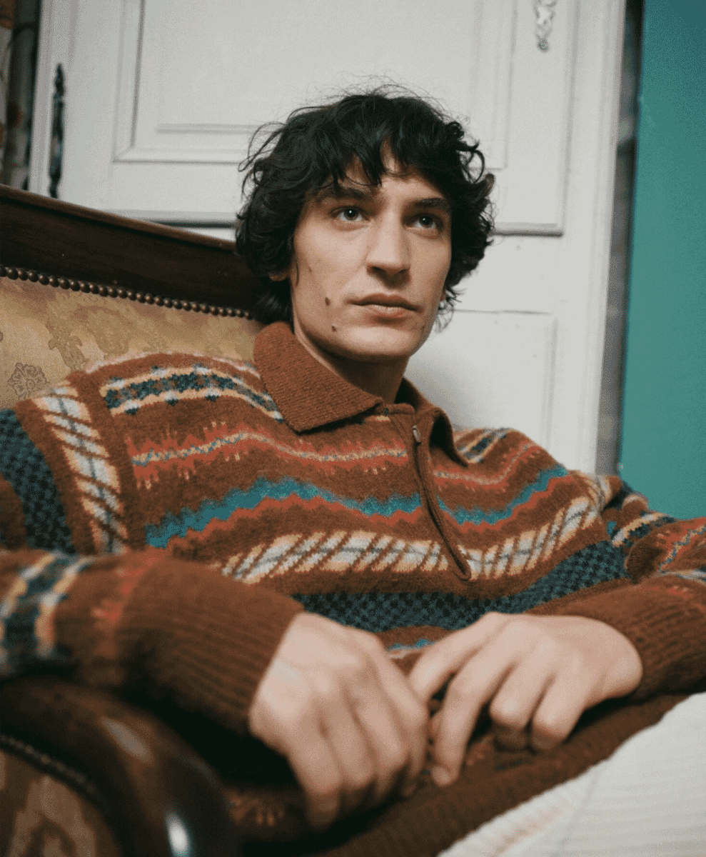 A man sitting on a vintage sofa wearing a brown and multi-colored patterned zip-neck sweater.