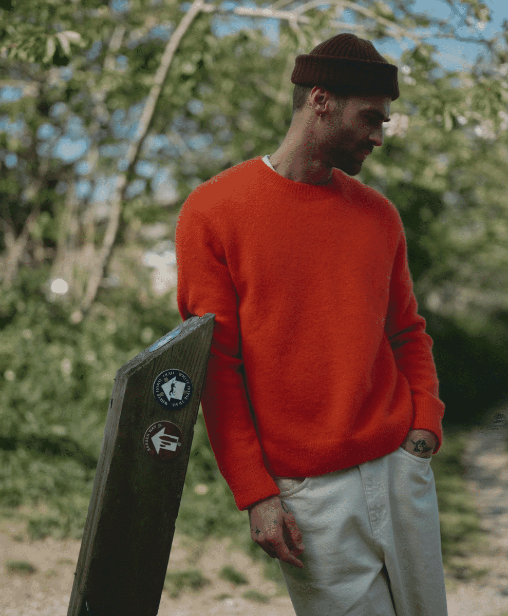 A man in an orange sweater and brown beanie standing in a sunlit forest.