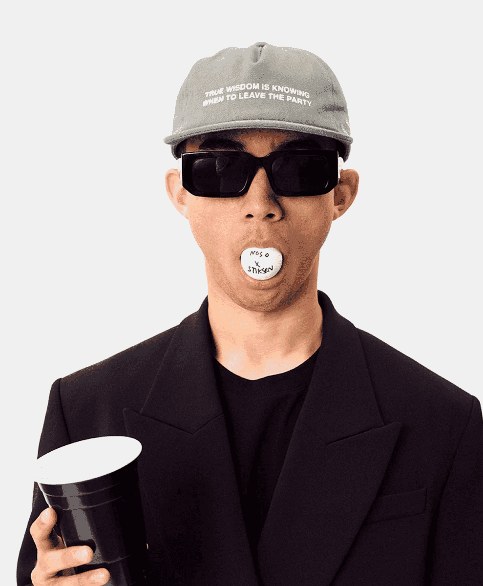 Man in black blazer and sunglasses wearing a 'TRUE WISDOM' cap.