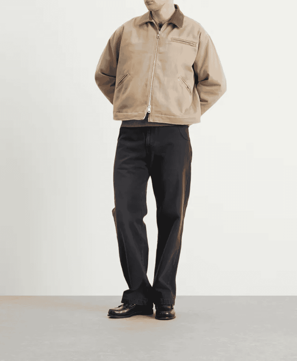 Man in a beige zip-front jacket and dark wide-leg trousers walking against a white background.