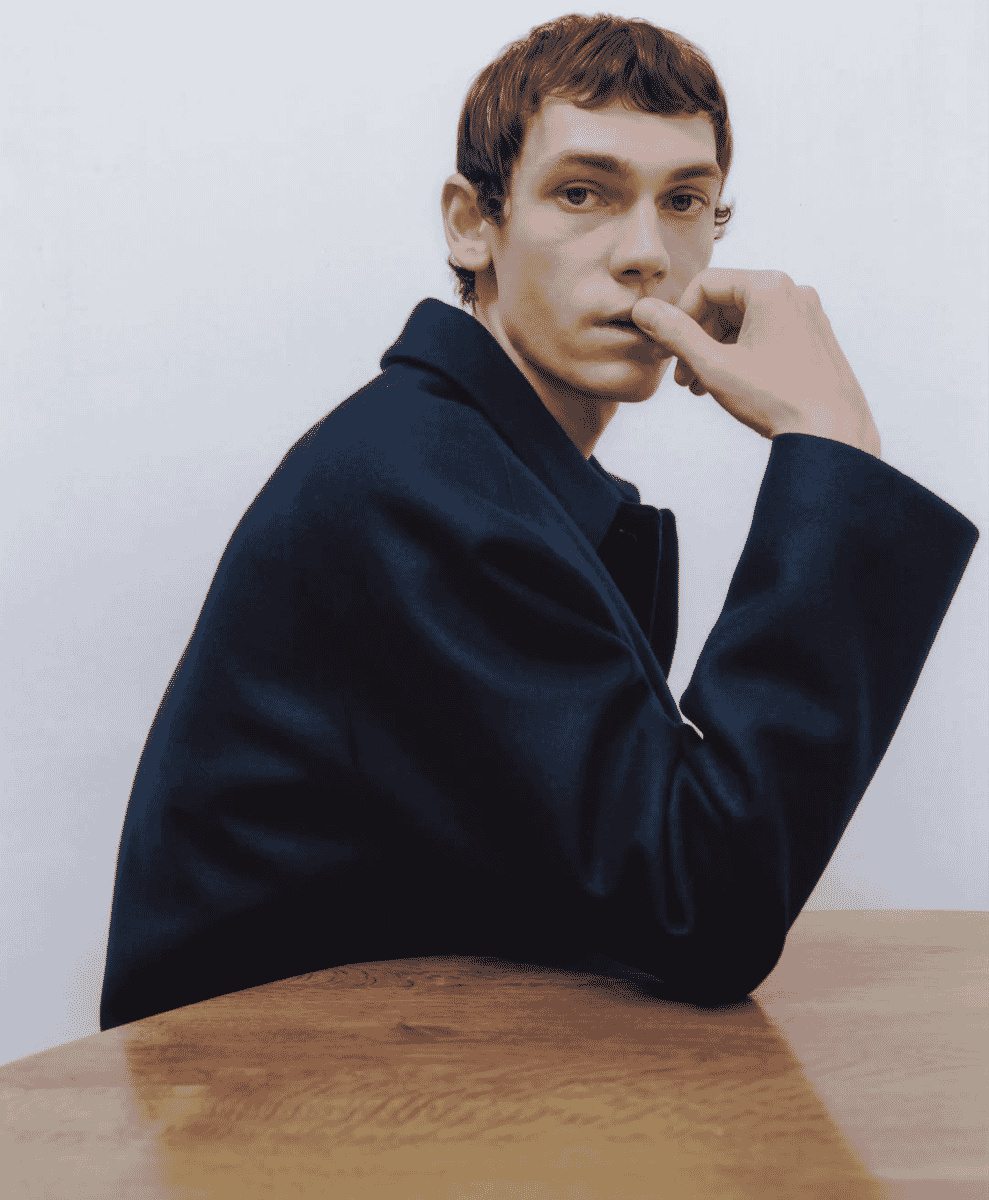 A man posing in a dark navy wool coat in a minimalist studio setting.