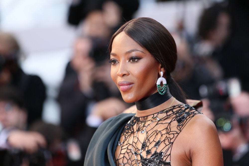 Naomi Campbell in a black beaded lace gown with dramatic green earrings on a red carpet.
