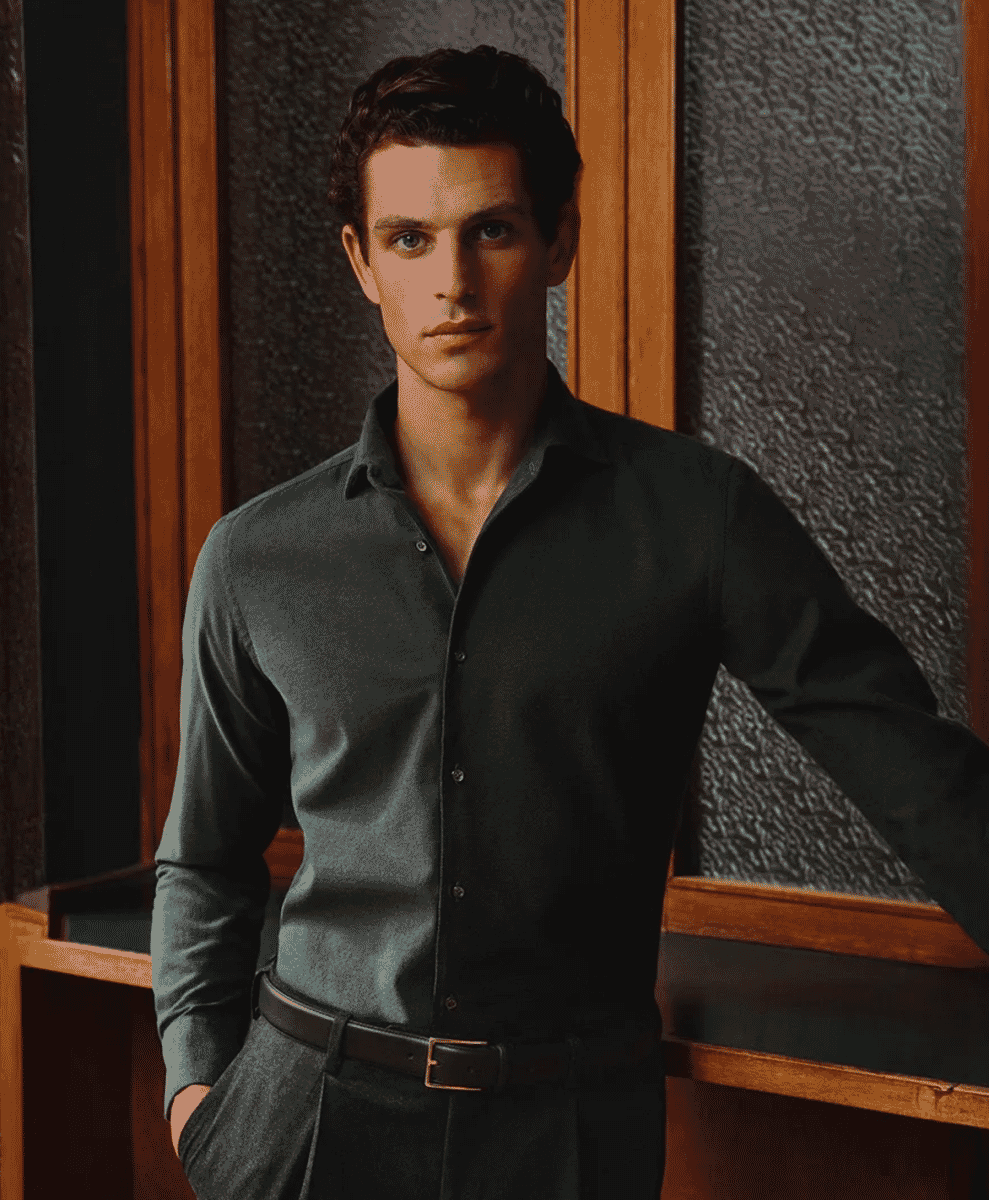 A man in a dark grey button-up shirt and matching trousers against a textured wall.