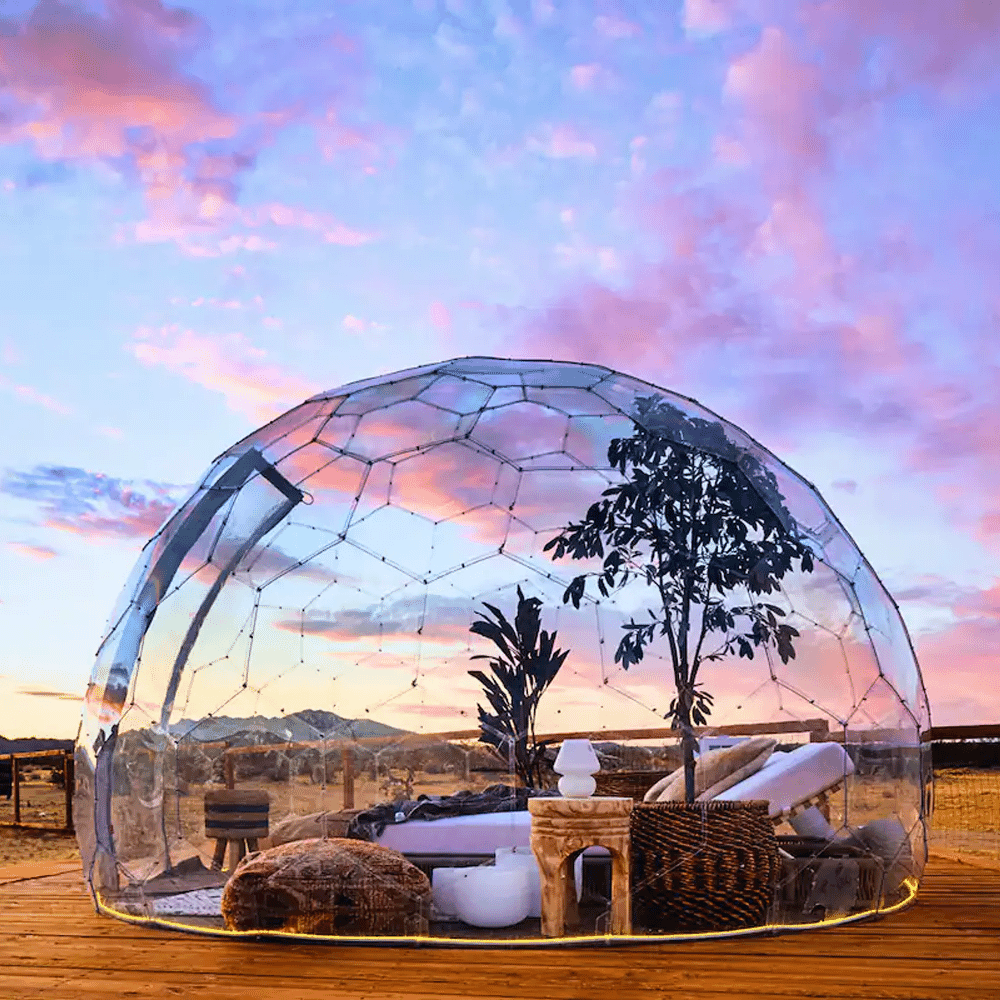 A transparent geodesic dome on a wooden deck at sunset with a view of distant mountains.