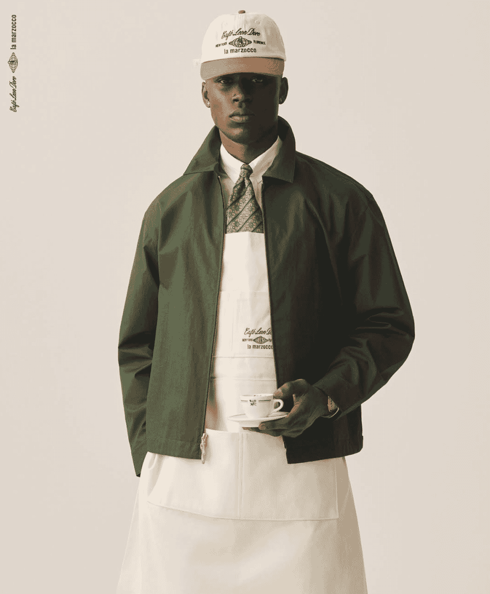 A model in a minimalist studio wearing an olive green jacket and a collaborative branded apron.