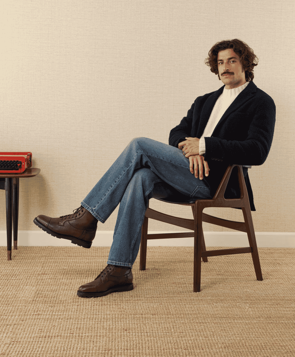 A man in a navy blazer, white turtleneck, jeans, and brown leather boots.