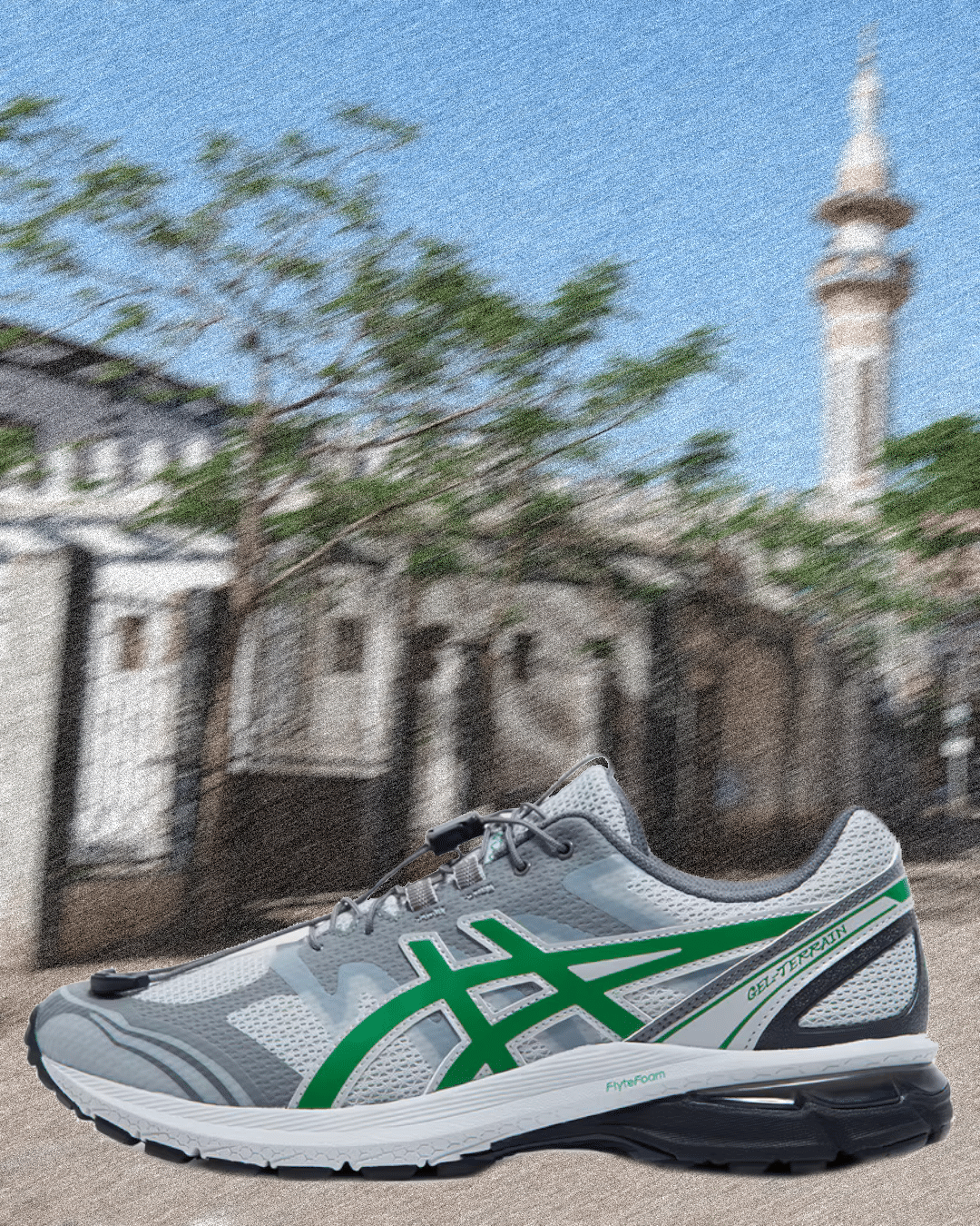 A gray and green Asics Gel-Terrain running shoe in front of a blurred mosque and traditional buildings.