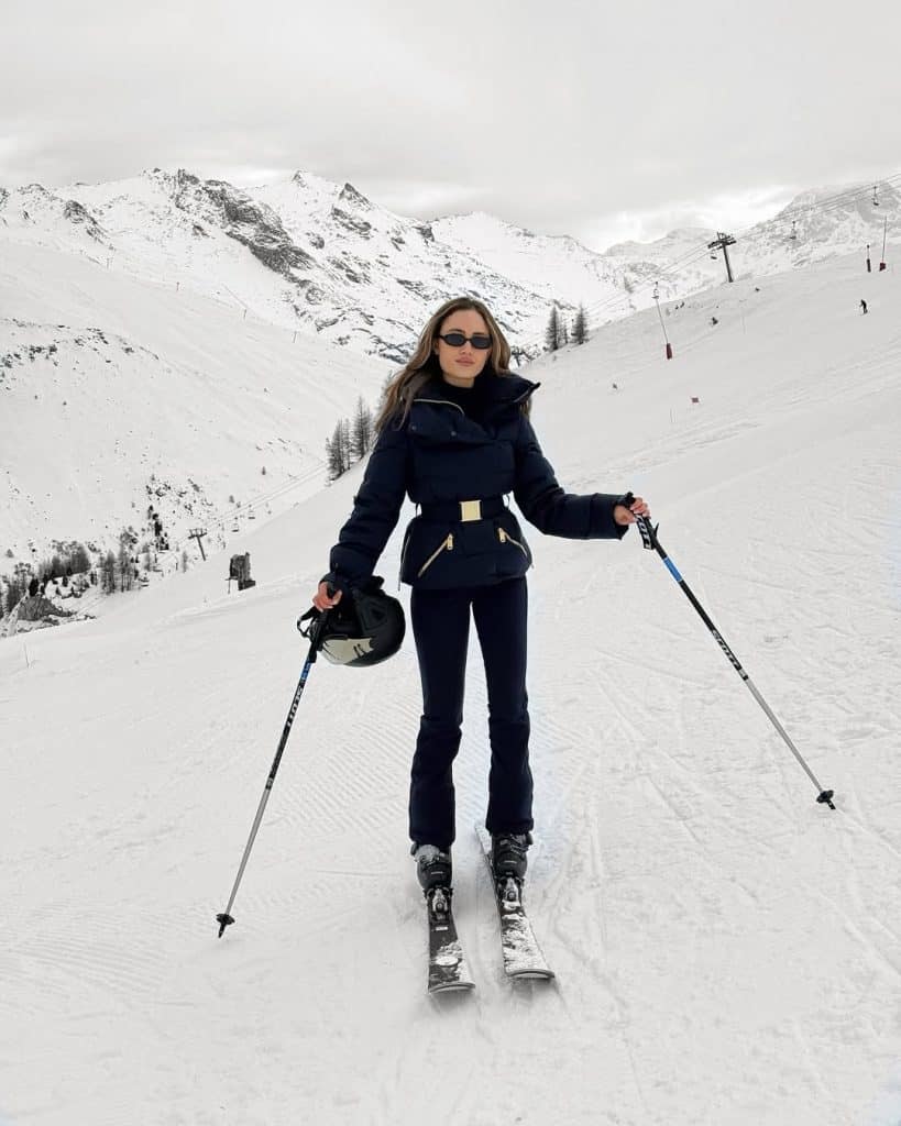 Woman in a navy blue ski jacket with gold hardware posing on a snowy slope.