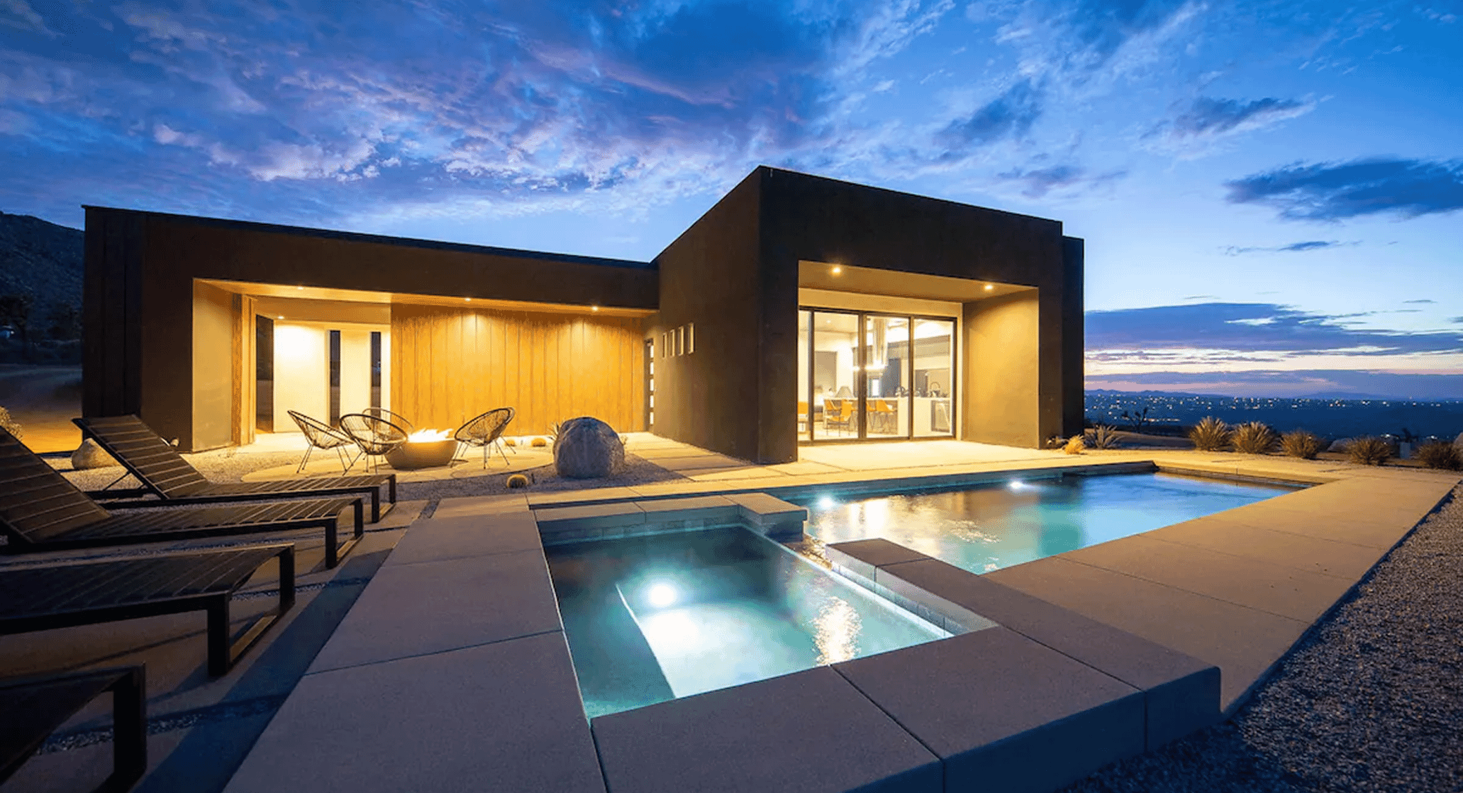 Modern minimalist house at dusk with illuminated pool and patio furniture against a desert landscape.