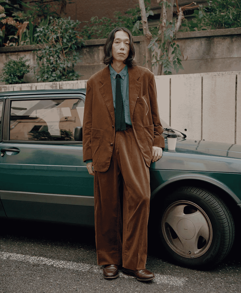 A man wearing a brown corduroy suit standing next to a vintage green car.