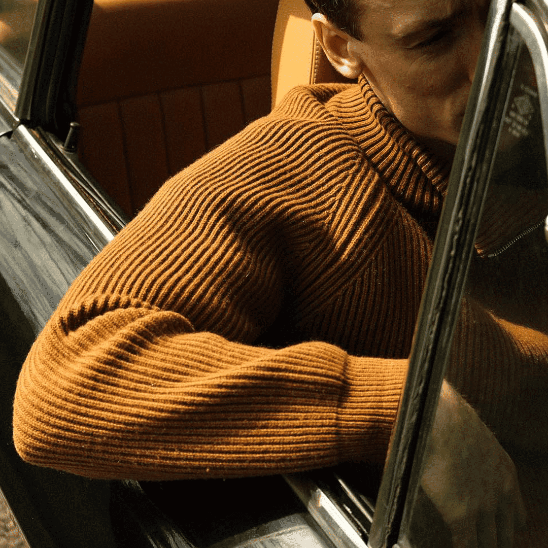 Man in a brown ribbed turtleneck sweater leaning out of a vintage car in warm sunlight.