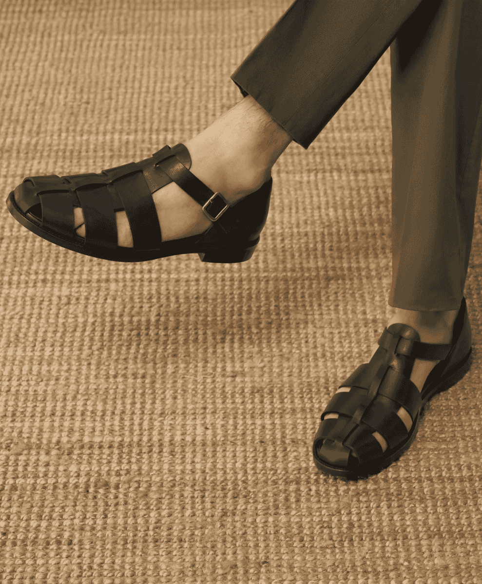 Black leather gladiator sandals paired with dark tailored trousers on a beige rug.