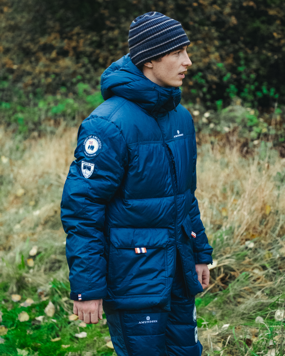 A man in a navy blue Amundsen puffer jacket with patches and matching beanie in an autumnal field.