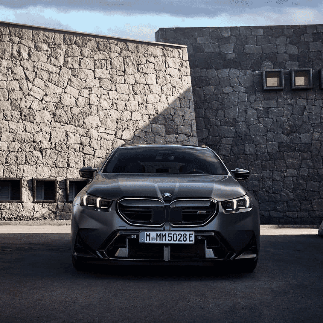 Frontal view of a gray BMW M5 Touring parked against a modern stone wall, highlighting its LED headlights and wide front fenders.