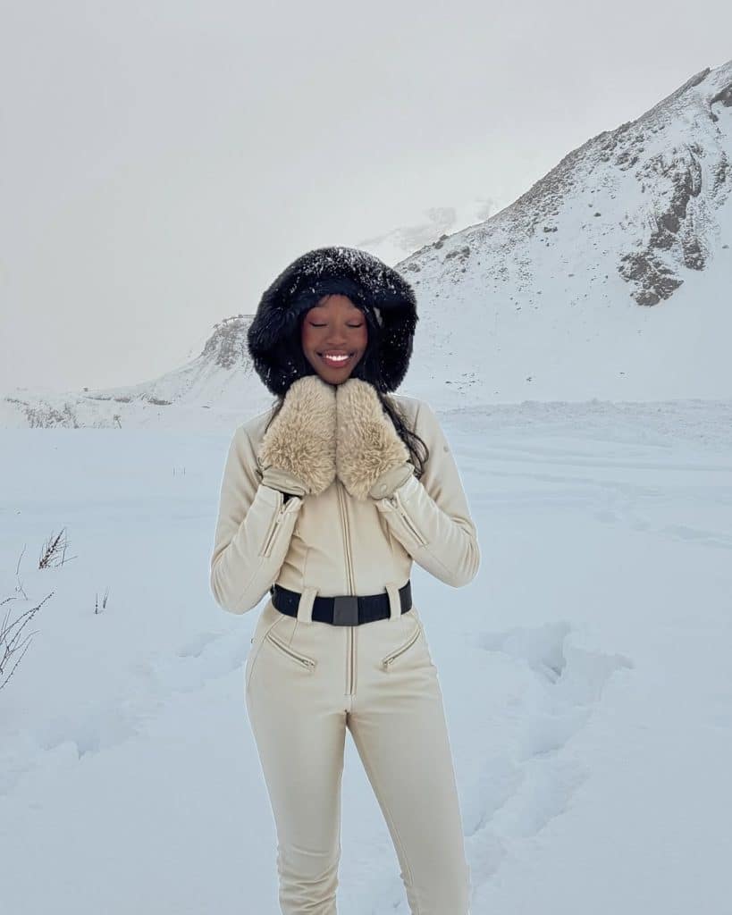 Woman in a cream-colored ski suit with a black fur-lined hood in a snowy landscape.