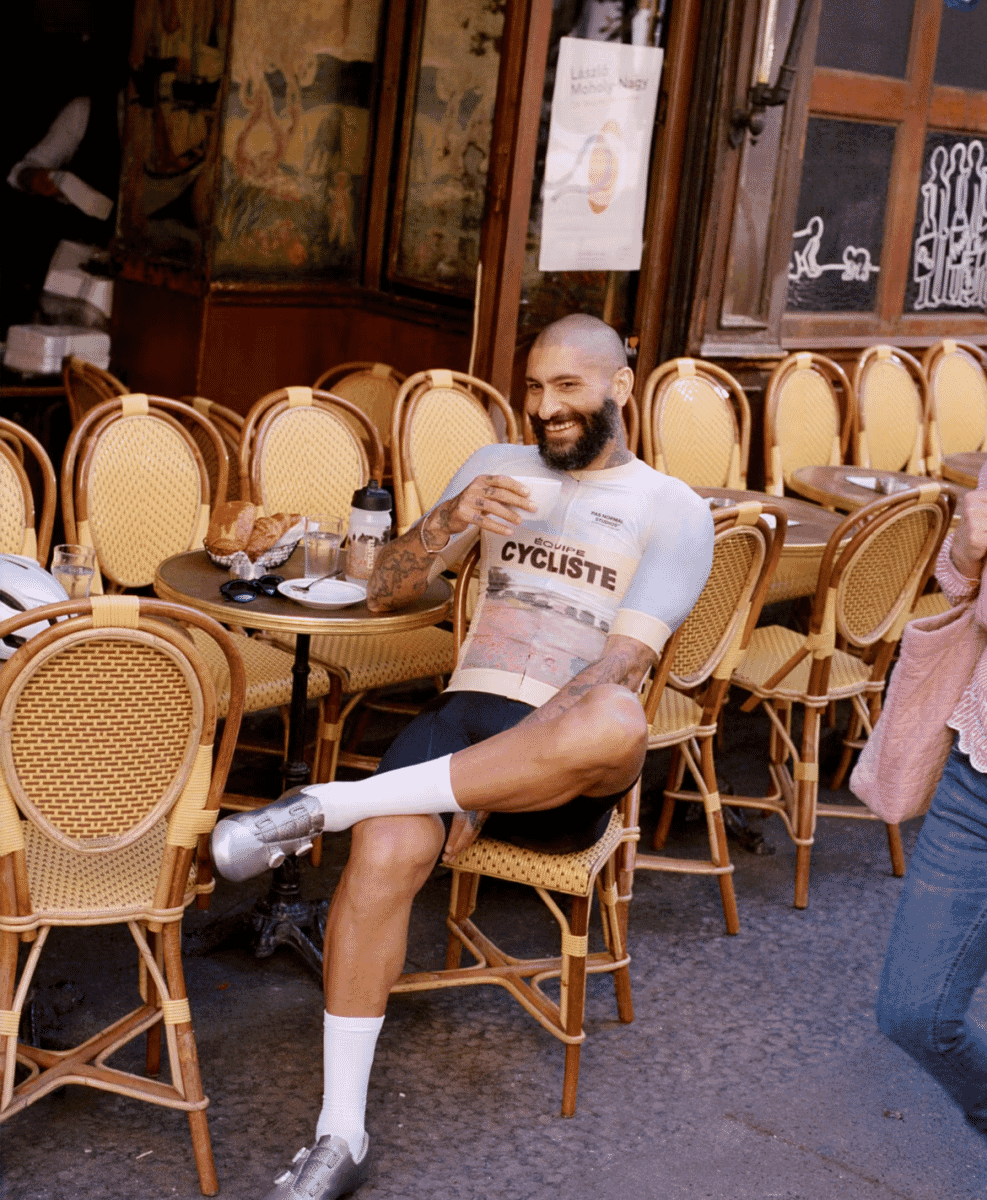 A bearded man in a white cycling jersey sitting at a café table with a cup of coffee.