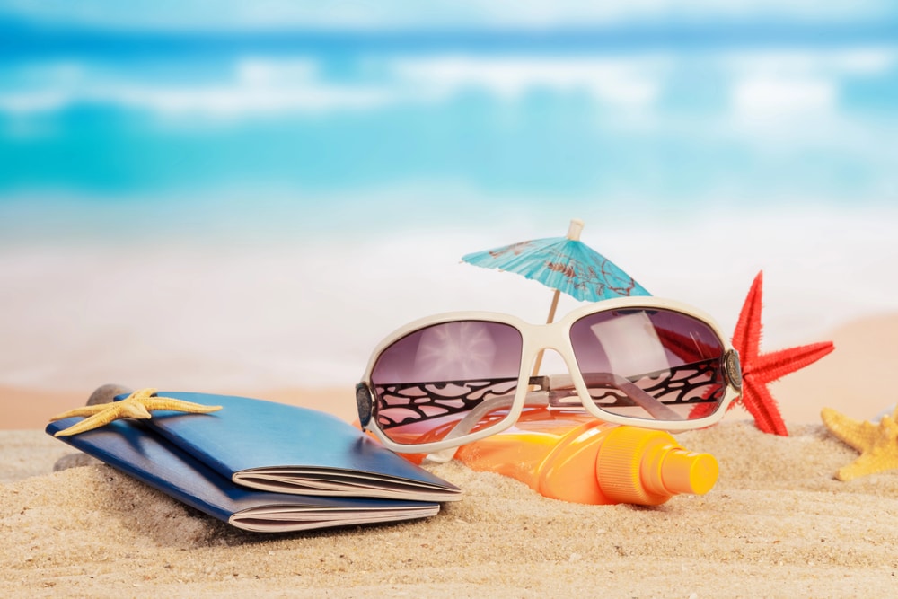 Sunscreen bottle and white-framed sunglasses arranged on beach sand.