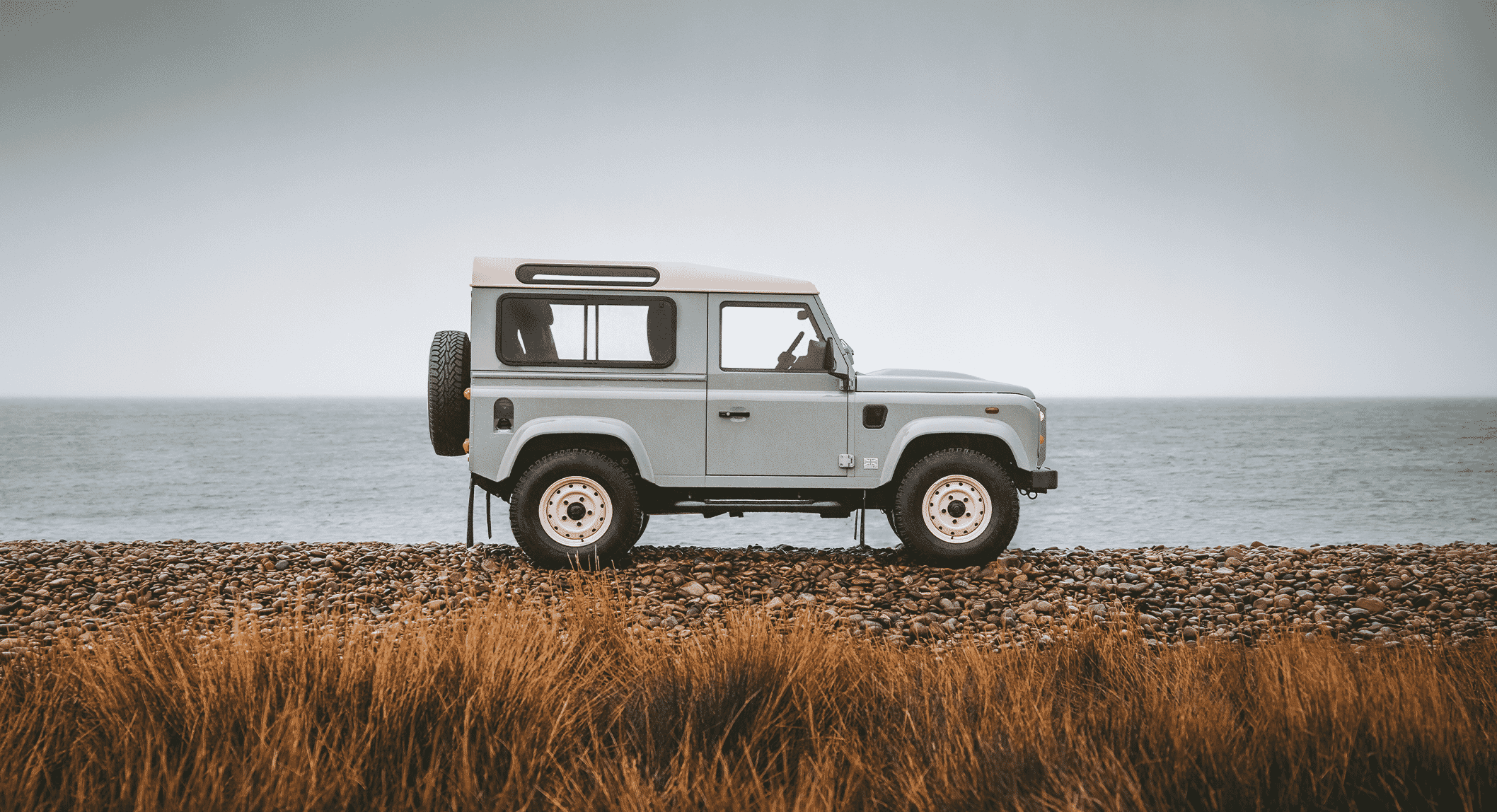 A light gray classic Land Rover Defender on an Islay pebble beach with tall grass in the foreground.