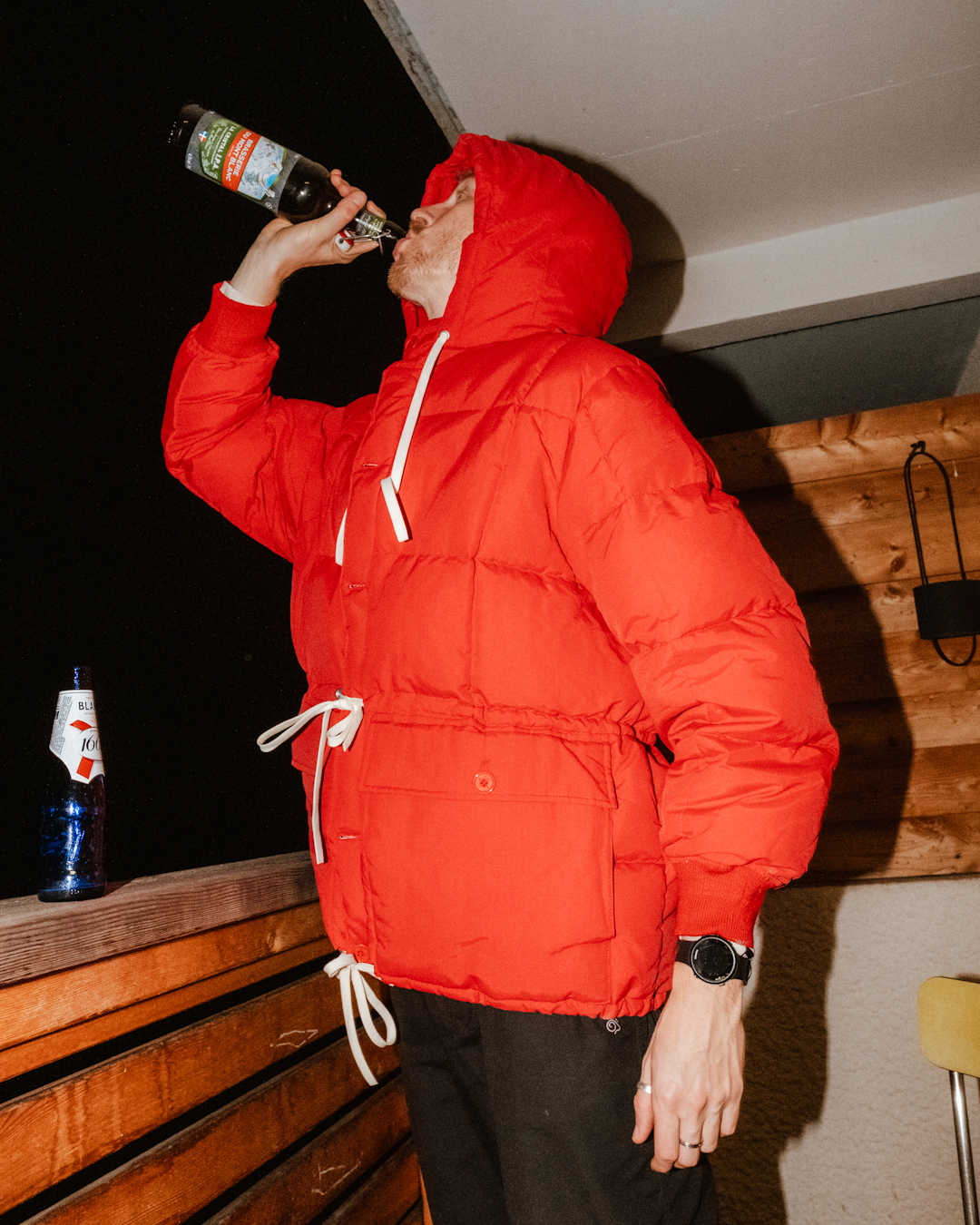 A man in a vibrant red hooded puffer jacket drinking from a beer bottle on a wooden balcony at night.