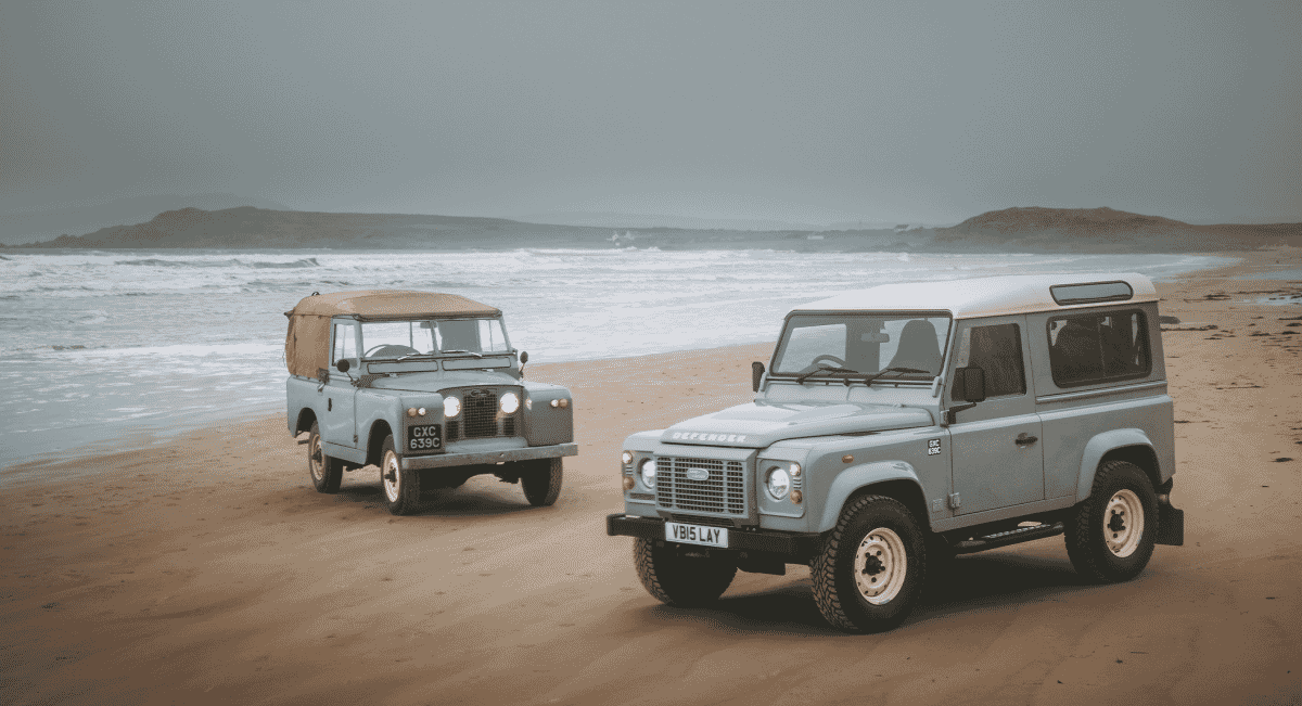 Two classic Land Rover Defenders in Heritage Grey parked on a sandy beach near the waves.