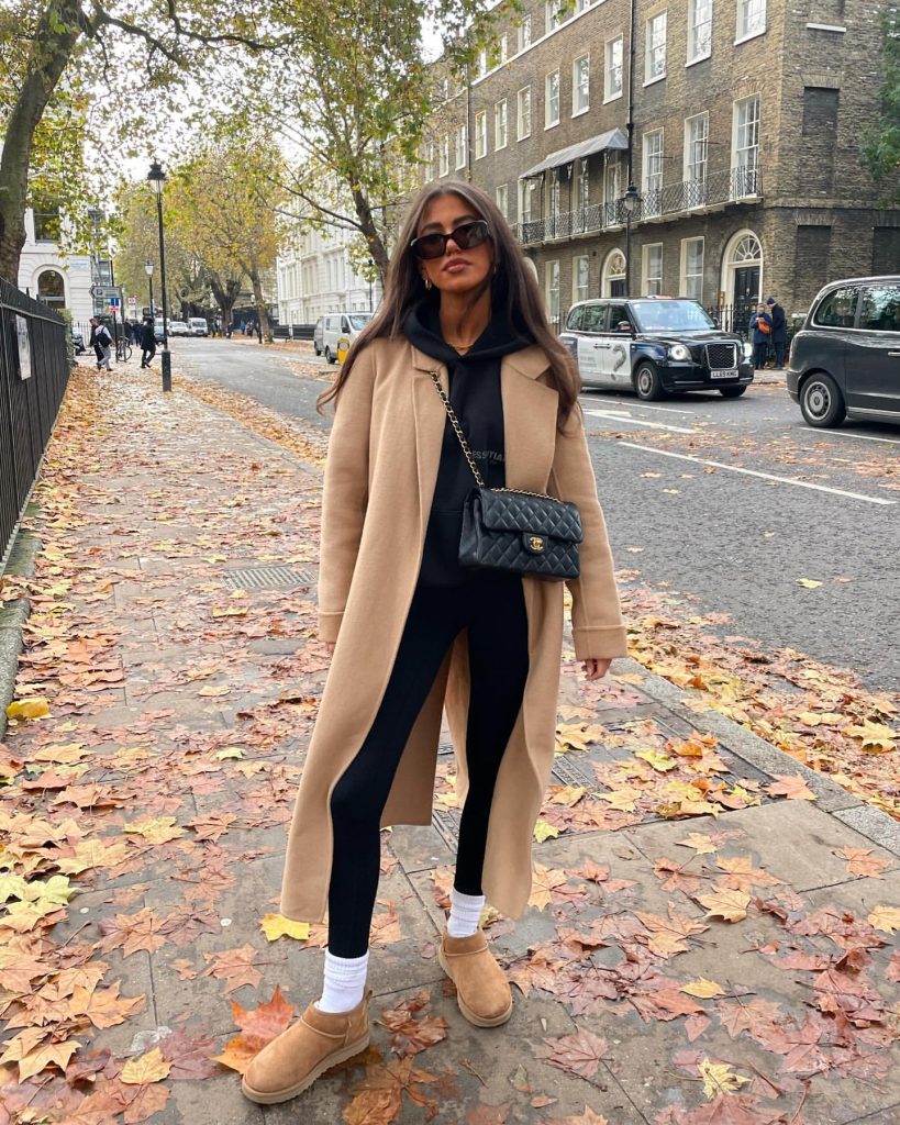 Woman in a camel coat, black hoodie, and black leggings walking on a London sidewalk.