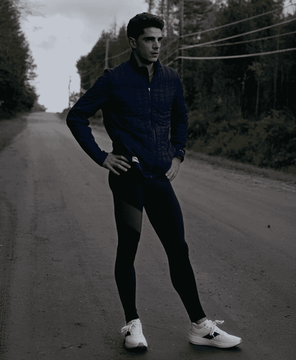 A man in a navy quilted jacket and black compression leggings standing on a rural road.