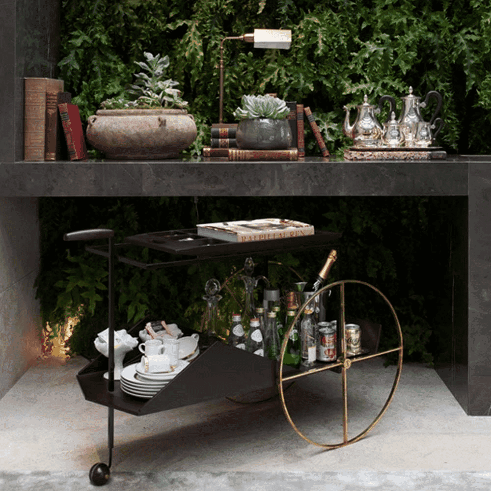 A black and gold bar cart styled with vintage books, silver teapots, and lush greenery.