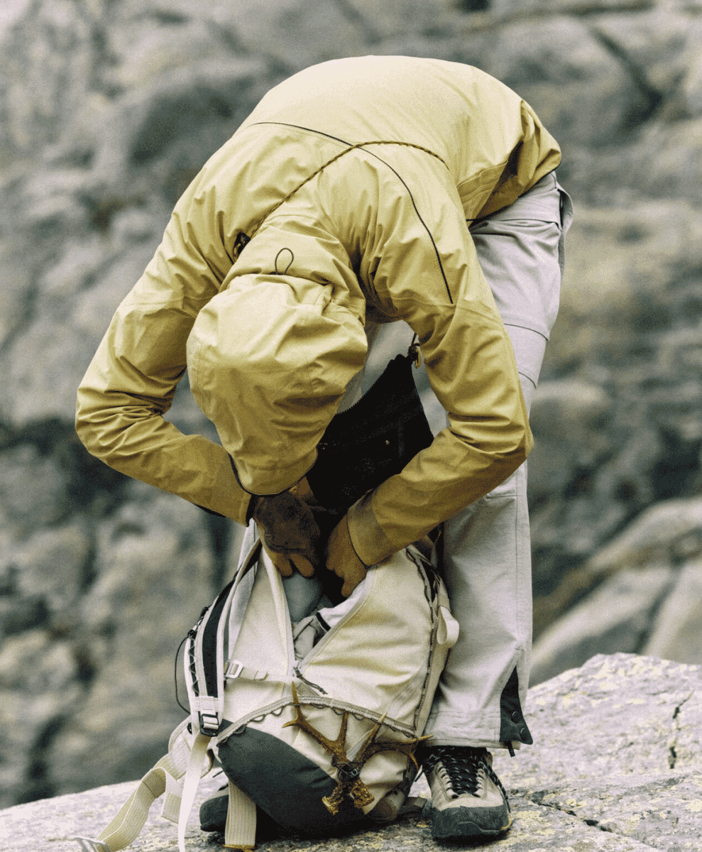 A hiker in a tan jacket bending over to organize gear inside a white Ull technical backpack on a rocky ledge.