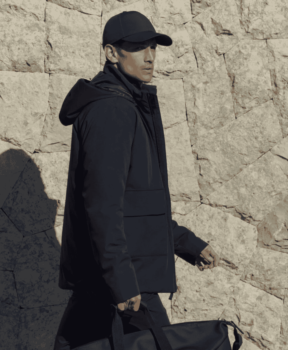 Man walking by a stone wall in a minimalist black hooded jacket and matching accessories.