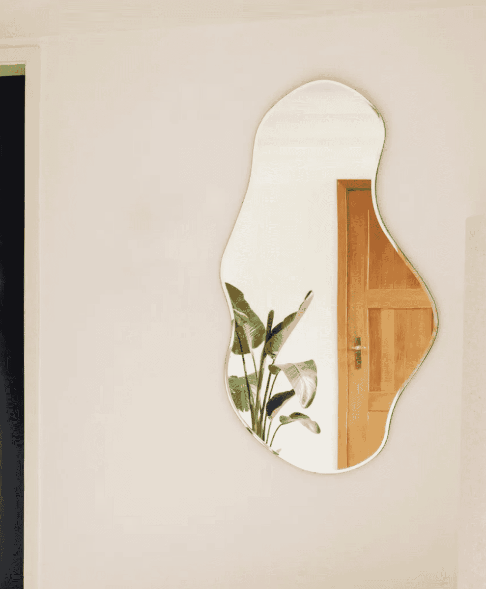 Abstract frameless mirror on a cream wall reflecting green plants and a wooden door.