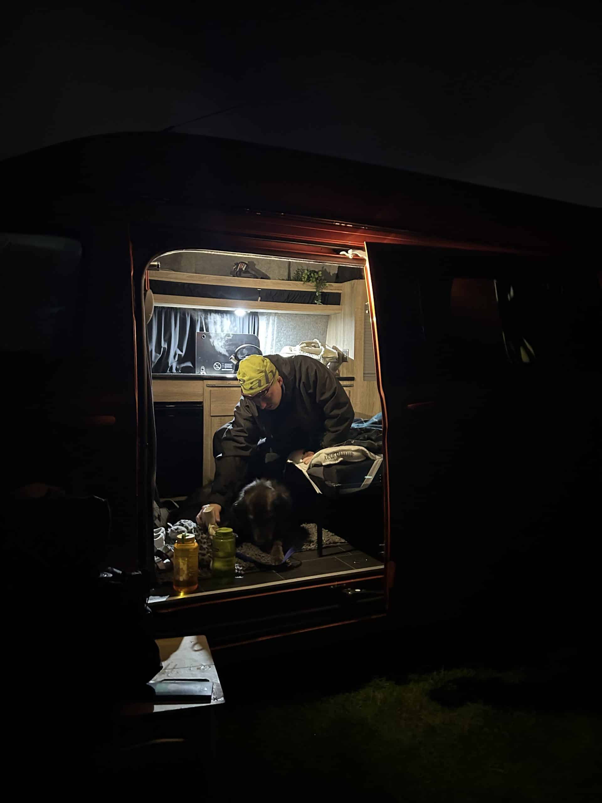A person in a yellow beanie and technical jacket inside a warmly lit camper van at night.